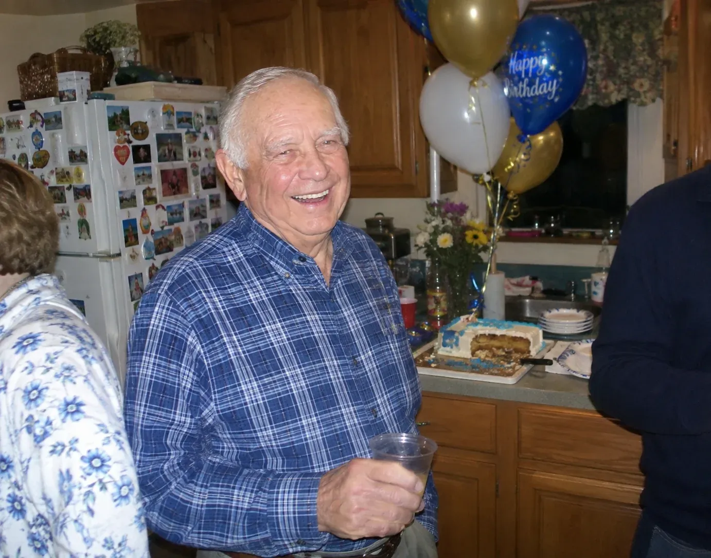 An ordinary phone snapshot of a laughing older man in a plaid shirt at a birthday party, in a crowded kitchen with balloons, a cake and other guests around him.